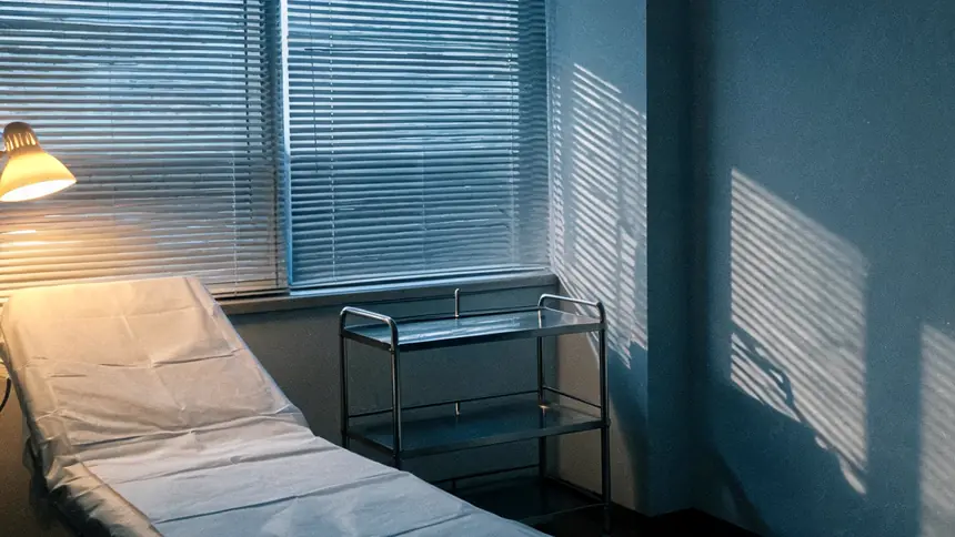 An empty consultation room at dusk, examination couch and instrument trolley under blinds.