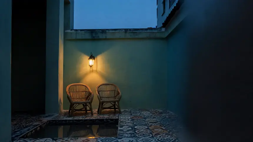 The airwell courtyard of a restored shophouse guesthouse at dusk, two rattan chairs under a single lantern.