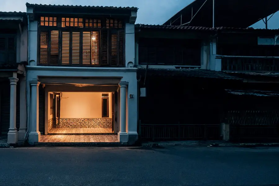 One Penang shophouse unit open and warmly lit at dusk, its five-foot way tiled, the units either side shuttered and dark.