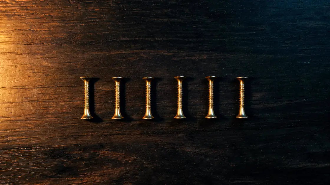 Six identical brass screws laid out in a row on a dark workbench, evenly spaced, each catching the same warm light.
